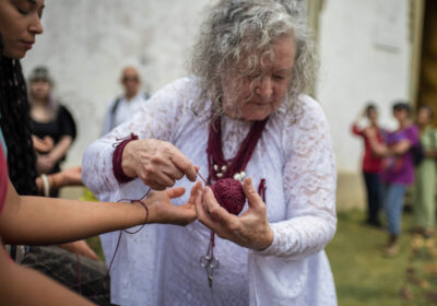 Fotografia em plano médio da artista Cecilia Stelini, uma mulher de cabelos brancos e cacheados, vestindo uma túnica branca rendada de mangas longas. Ela segura com as duas mãos um novelo de linha tom bordô e atesta com cuidado um fio ao redor do pulso de outra pessoa em primeiro plano. Ela carrega colares de fios escuros e uma pequena tesoura pendurada na gola. Ao fundo desfocado, pessoas observam a ação ritual ao ar livre diante de uma parede clara. Foto: Saulo Santiago.